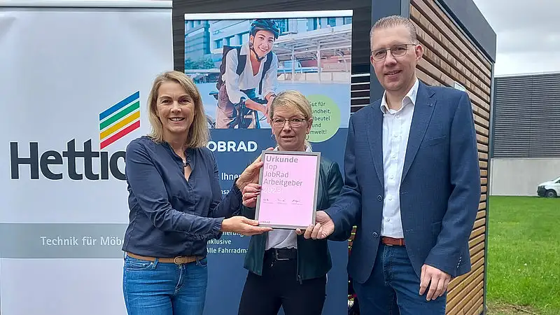 Monika Titze, von der Hettich Gruppe, steht zusammen mit einer Frau und einem Mann vor einem Poster mit dem Schriftzug Hettich und dem Hettich Logo. Sie alle tragen blaue Geschäftskleidung Monika Titze, von der Hettich Gruppe, steht zusammen mit einer Frau und einem Mann vor einem Poster mit dem Schriftzug Hettich und dem Hettich Logo. Sie alle tragen blaue Geschäftskleidung