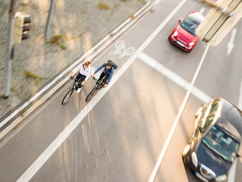 Zwei Fahrradfahrer:innen im Straßenverkehr mit Autos, von schräg vorne/oben fotografiert
